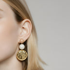Close-up of an ear wearing a gold earring with a round design, pearl, and beige bead against a neutral background.